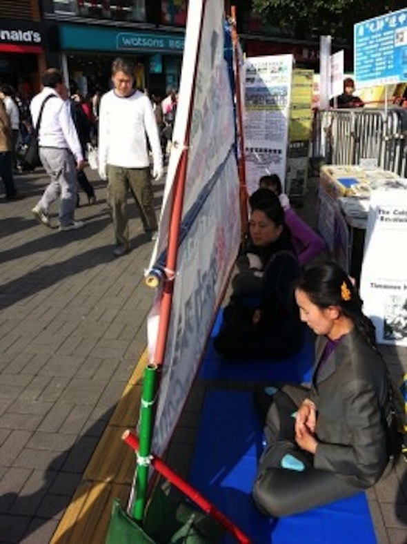 Falun Gong practitioners are seen meditating at their information site at Star Ferry in Hong Kong on Feb. 13, 2013. A rack of banners subsequently erected by the Hong Kong Youth Care Association blocks passerby from seeing the practitioners' site. The raised tile at the base of the Association's banners is a pathway meant for the blind. (Epoch Times)
